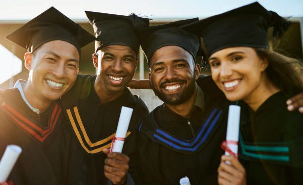 Portrait of diverse group of graduates holding degree and diploma during graduation ceremony outside on university campus. Smiling and happy friends standing close together at college. Gown and cap